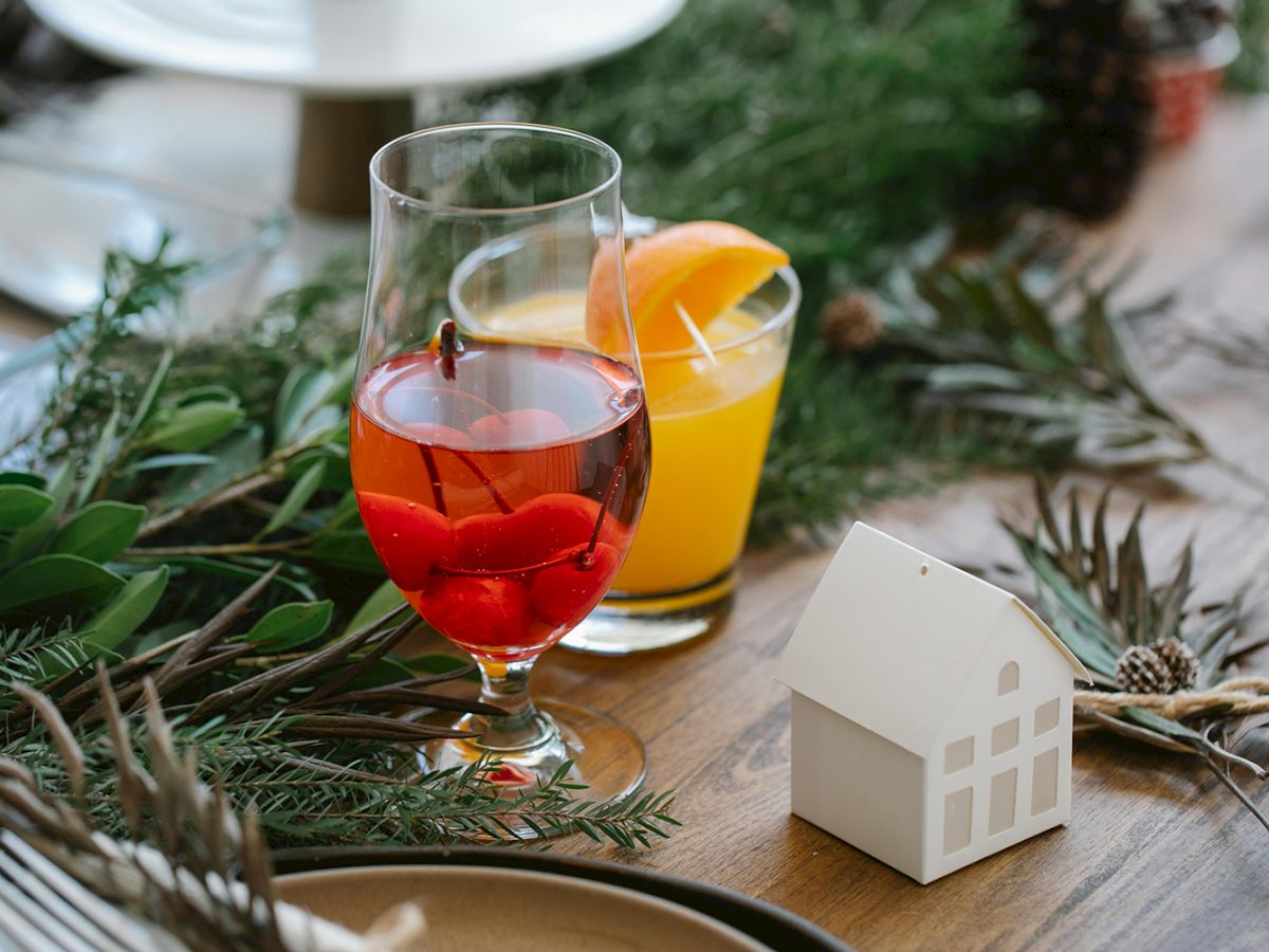 A festive table setting features two drinks—a red one with berries and an orange one with a slice—amid greenery, pinecones, and a small house decoration.