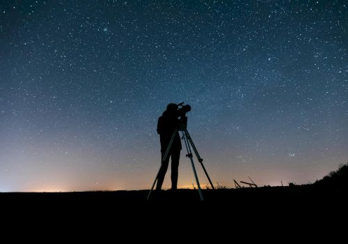 A person is using a telescope under a clear, starry night sky, silhouetted against the horizon with city lights faintly glowing in the distance.