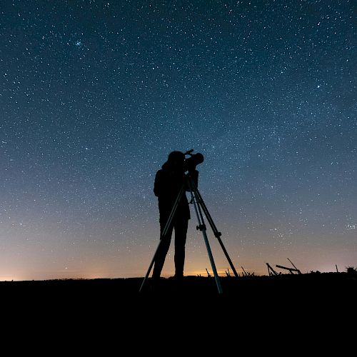 A person is using a telescope under a clear, starry night sky, silhouetted against the horizon with city lights faintly glowing in the distance.