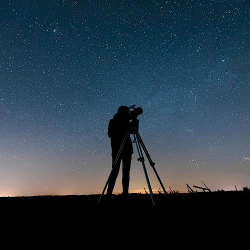 A person is using a telescope under a clear, starry night sky, silhouetted against the horizon with city lights faintly glowing in the distance.