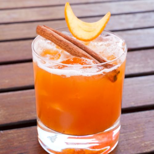 A glass of an orange-colored cocktail topped with a cinnamon stick and a slice of citrus peel, placed on a wooden table.