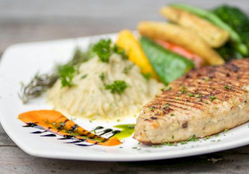 A plated dish featuring a grilled fish fillet, rice topped with herbs, baby corn, and other vegetables on a white plate garnished with sauce.