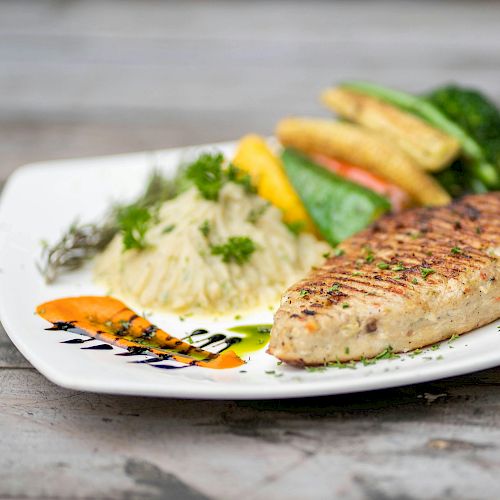 A plated dish featuring a grilled fish fillet, rice topped with herbs, baby corn, and other vegetables on a white plate garnished with sauce.