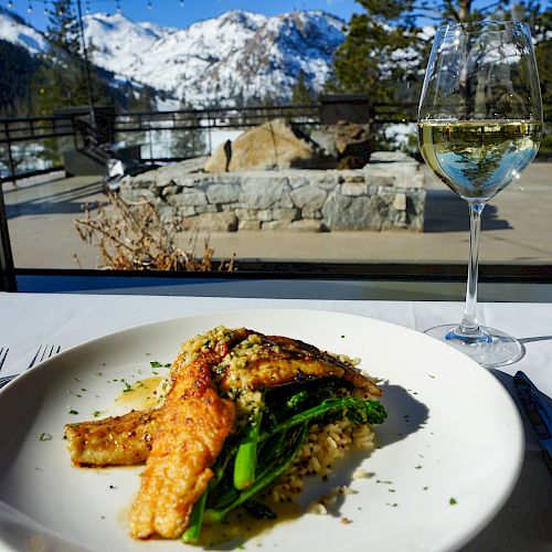 A beautifully plated dish with vegetables, paired with a glass of white wine, set against a stunning snowy mountain view from a window.