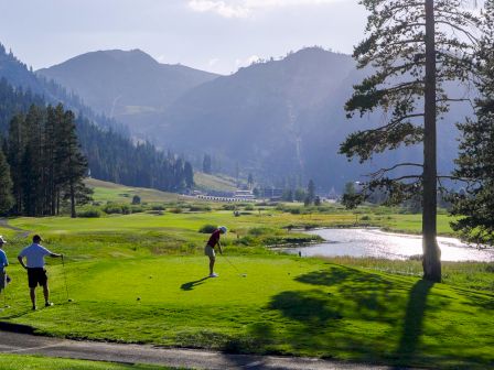 Two people are golfing on a lush green course surrounded by mountains and trees, with a golf cart nearby under a clear sky.