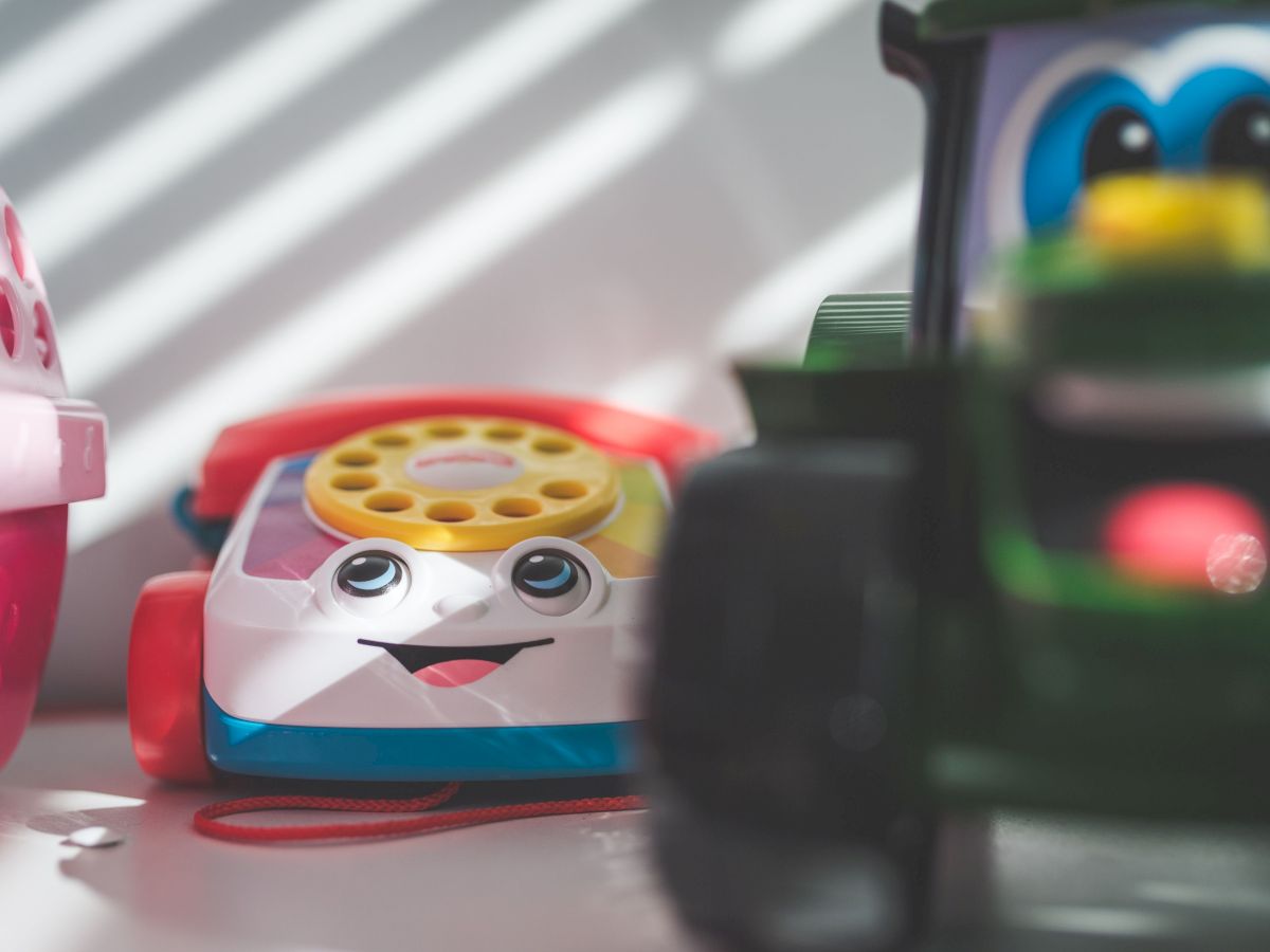 A colorful smiling toy vehicle/robot with big eyes and a yellow dial sits amid other play pieces in a blurred background.