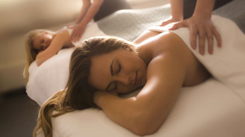 Two people are receiving massages on white beds, with hands visible applying pressure on their backs.