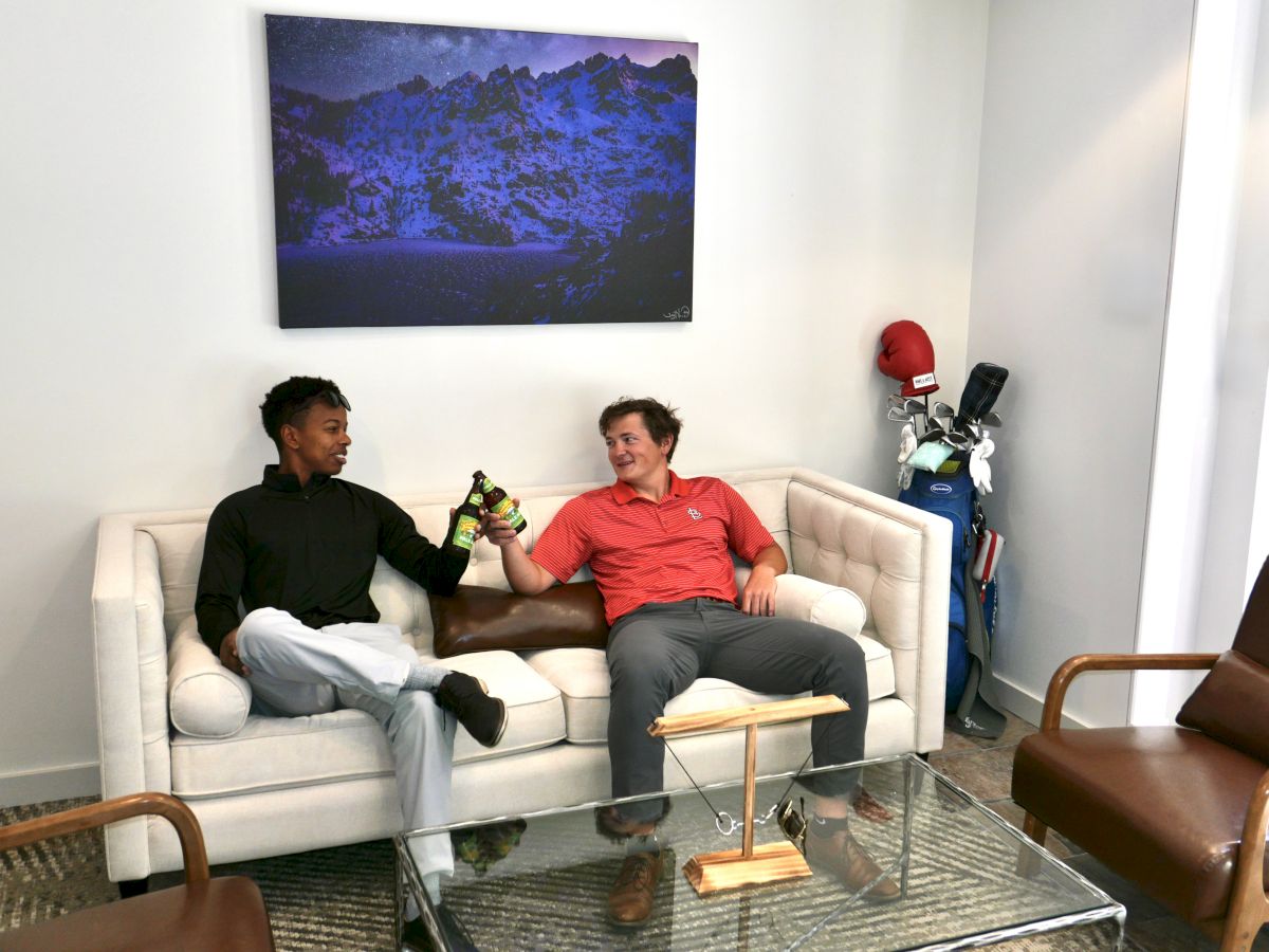 Two men lounge on a white couch chatting and holding beer bottles while a red boxing-glove figure and golf gear stand nearby.