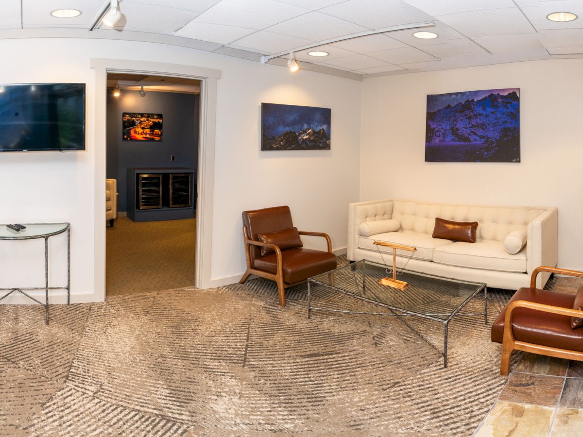 A modern office lounge with couches, chairs, a glass coffee table, wall TVs, and mountain artwork under ceiling lights.