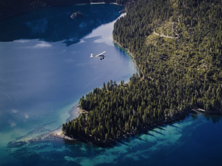 Aerial view shows a seaplane over a turquoise lake beside dense pine forest and rocky shores.