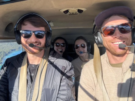 Four people in a plane cockpit wearing headsets and sunglasses, smiling with microphones as they fly over scenery.