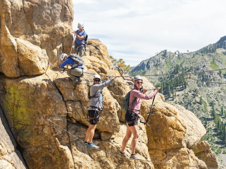 Hikers in helmets climb rugged rocky cliffs, taking photos against a mountain valley backdrop.