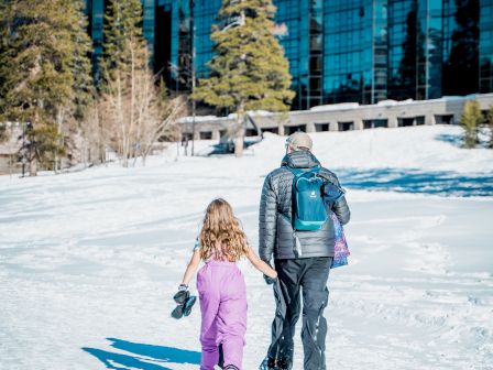 A snowy day as an adult and child walk across the snow toward a modern building in a pine-lined mountain area.