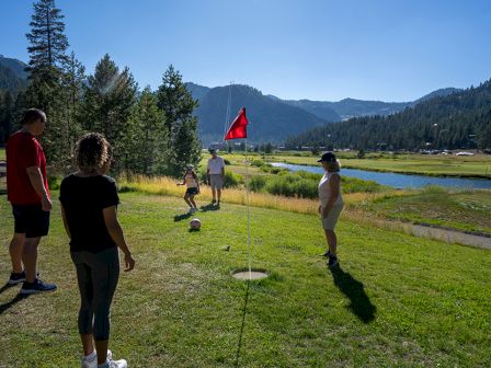 A group of people walk on a grassy course near a lake, with a red flag marking a disc-golf-style hole.