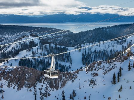 A snowy mountain cable car cabin hangs above pine forests and a frozen valley with a wide lake under dramatic clouds.