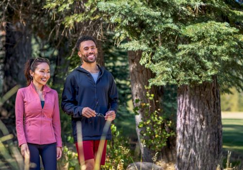 Two people are walking and smiling in a forested area, surrounded by trees and greenery.