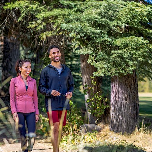 Two people are walking and smiling in a forested area, surrounded by trees and greenery.