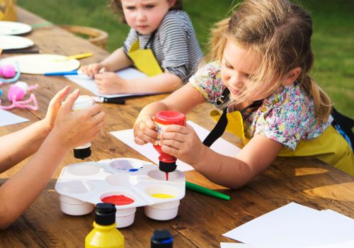 Children are engaged in an outdoor art activity, using paints and paper, with focus on creativity and fun.