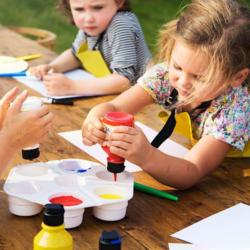 Children are engaged in an outdoor art activity, using paints and paper, with focus on creativity and fun.