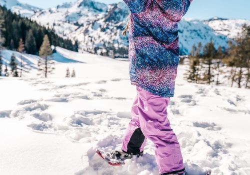 A person in colorful winter clothing is snowshoeing in a snowy mountain landscape with arms raised, enjoying the sunny day.