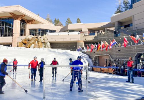 People are playing hockey on an outdoor ice rink near a building, with flags displayed above.