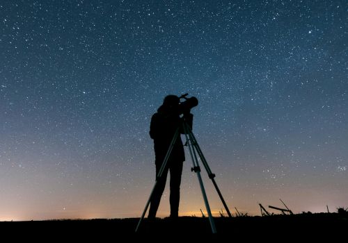 A person is silhouetted against a starry night sky, looking through a telescope on a tripod, capturing the beauty of the cosmos.