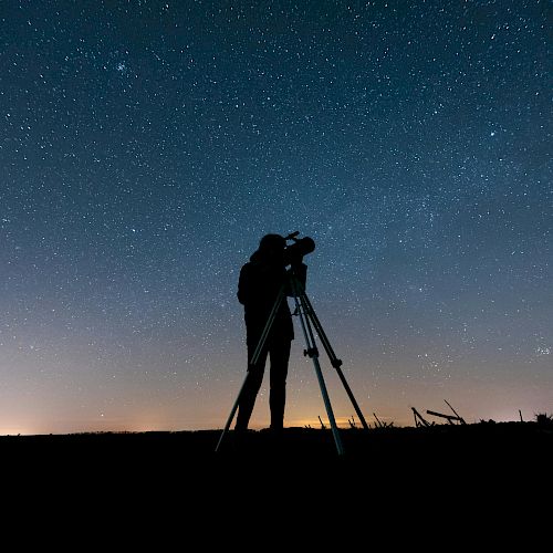 A person is silhouetted against a starry night sky, looking through a telescope on a tripod, capturing the beauty of the cosmos.