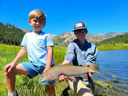 Two boys sit by a lake, smiling while holding a large trout in sunny mountains.