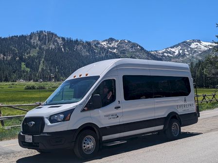 A white Everline van drives on a rural road with snowcapped mountains and green fields under a clear blue sky.