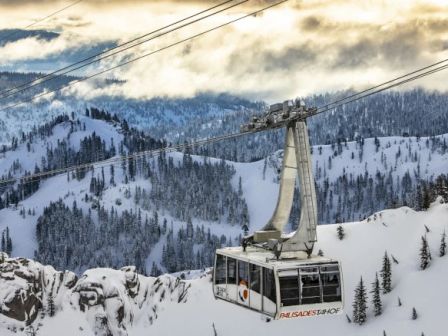 A cable car traverses snow-covered mountains with a cloudy sky above, surrounded by trees and rugged terrain.