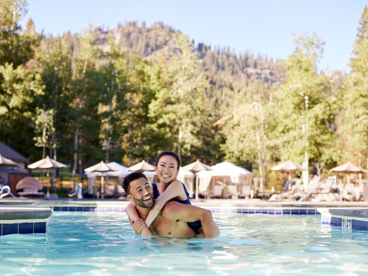 A couple enjoying a swim in an outdoor pool, surrounded by trees and mountains under a clear blue sky.