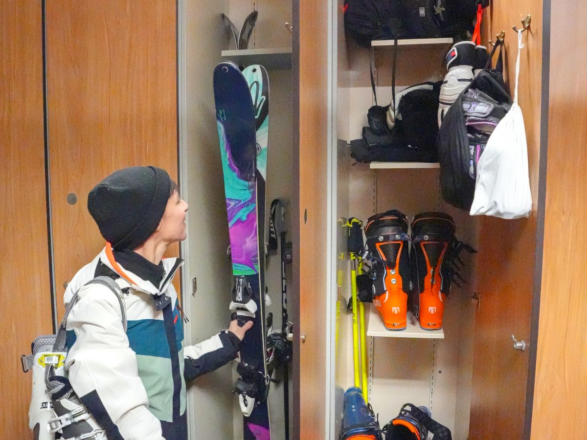 A person is standing in front of an open locker with skis, ski boots, helmets, and other skiing gear inside.