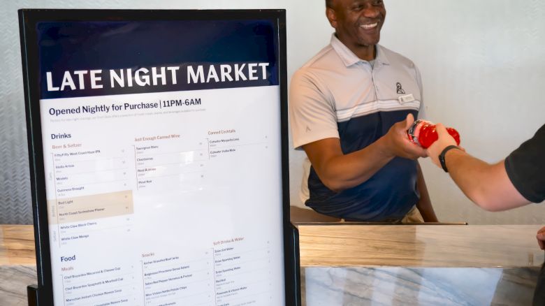 A server smiles while pouring a drink beside a &ldquo;Late Night Market&rdquo; menu board.