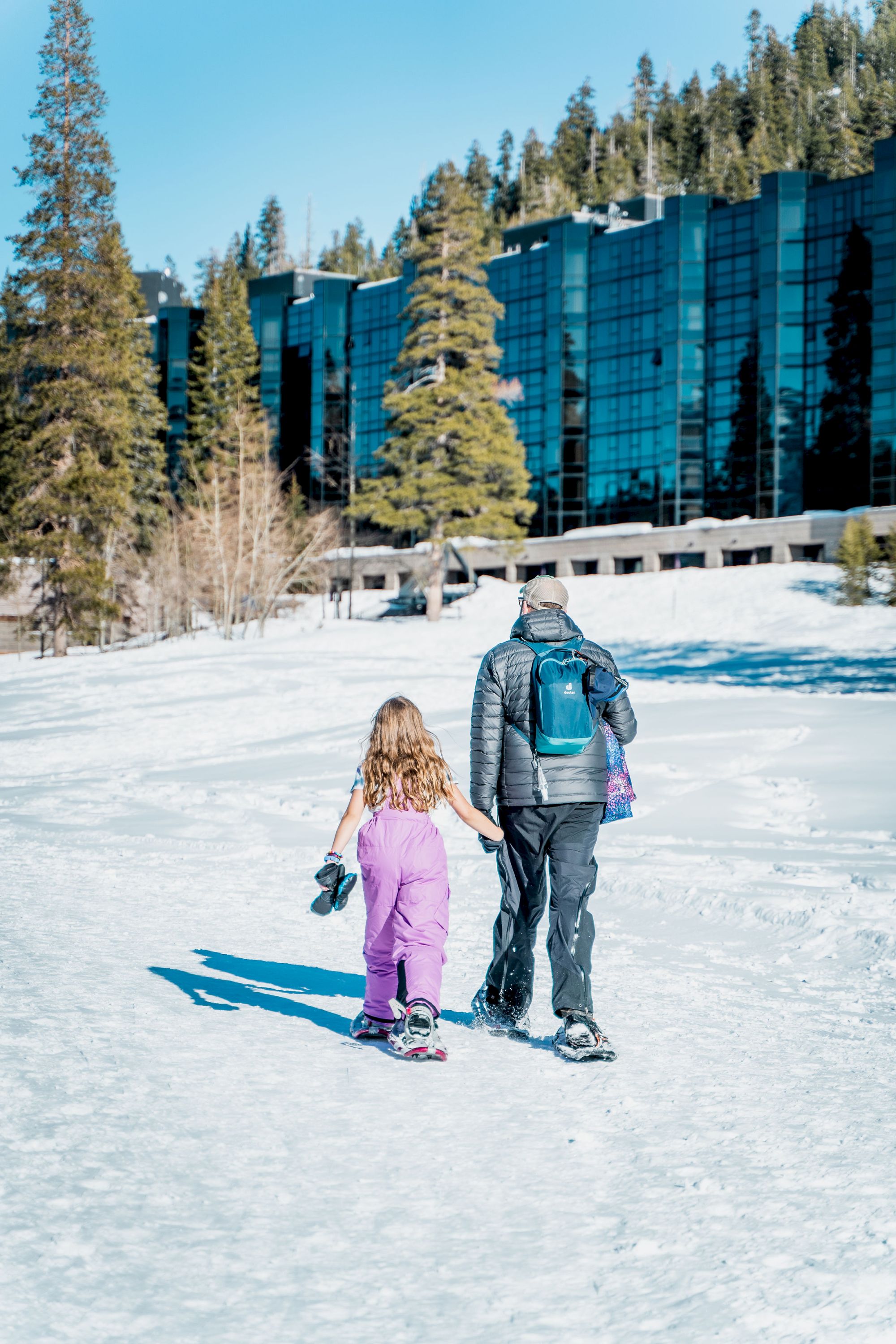 A person and a child in snow gear walk hand-in-hand on a snowy path toward a modern glass building surrounded by tall trees.