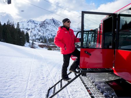 A person in winter gear steps into a red snow grooming vehicle on a snowy landscape, with mountains in the background.