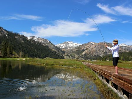 A person is fly fishing on a wooden dock beside a lake, with mountains and trees in the background under a blue sky with clouds.