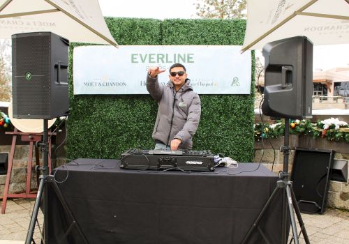 A DJ is performing outdoors, wearing sunglasses and a jacket, with music equipment and speakers, standing under umbrellas.