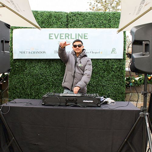 A DJ is performing outdoors, wearing sunglasses and a jacket, with music equipment and speakers, standing under umbrellas.