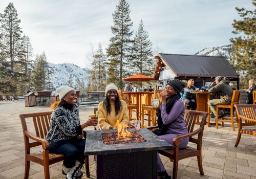 Three people sit around a fire pit outdoors, surrounded by trees and mountains. They are chatting and enjoying their time together.