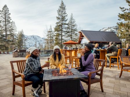 Three people are seated around a fire pit on a patio, surrounded by trees and snowy mountains, enjoying a relaxed outdoor setting.