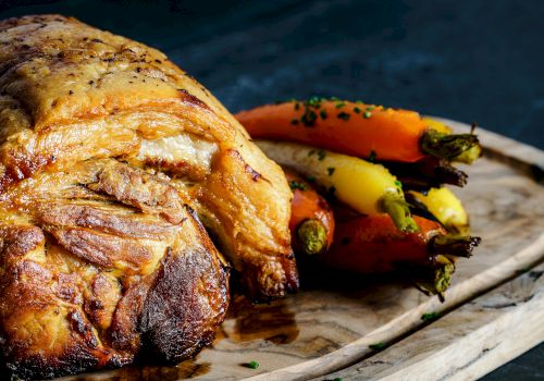 A roasted meat cut is displayed on a wooden board next to a bunch of colorful carrots, garnished with herbs.