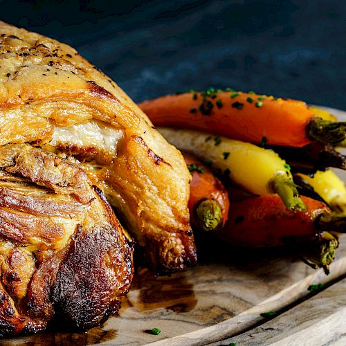 A roasted meat cut is displayed on a wooden board next to a bunch of colorful carrots, garnished with herbs.