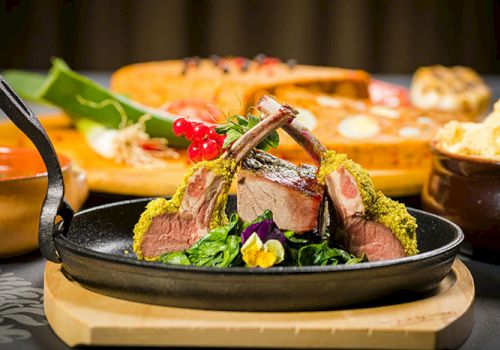 A plate with herb-crusted lamb chops on greens, garnished with berries and edible flowers; background shows bread and vegetables.