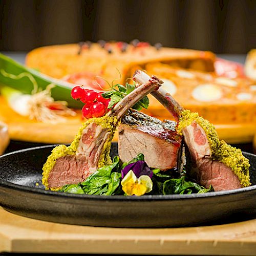 A plate with herb-crusted lamb chops on greens, garnished with berries and edible flowers; background shows bread and vegetables.