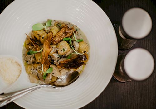 A dish of gnocchi with mushrooms and herbs is served, accompanied by grated cheese and two spice shakers on the side.