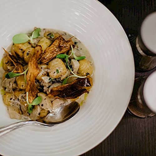 A dish of gnocchi with mushrooms and herbs is served, accompanied by grated cheese and two spice shakers on the side.