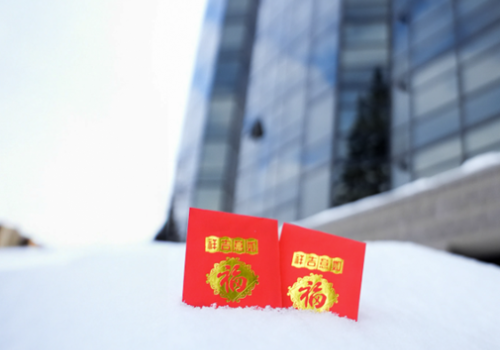 Two red envelopes with gold designs and Chinese characters are placed on a snow-covered surface in front of a glass building.