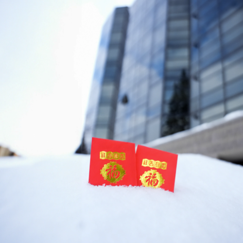 Two red envelopes with gold designs and Chinese characters are placed on a snow-covered surface in front of a glass building.