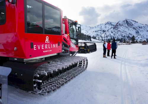 A red snow grooming vehicle on a snowy landscape with mountains and three people in winter clothing nearby.