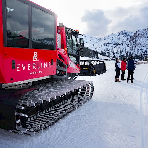 A red snow grooming vehicle on a snowy landscape with mountains and three people in winter clothing nearby.